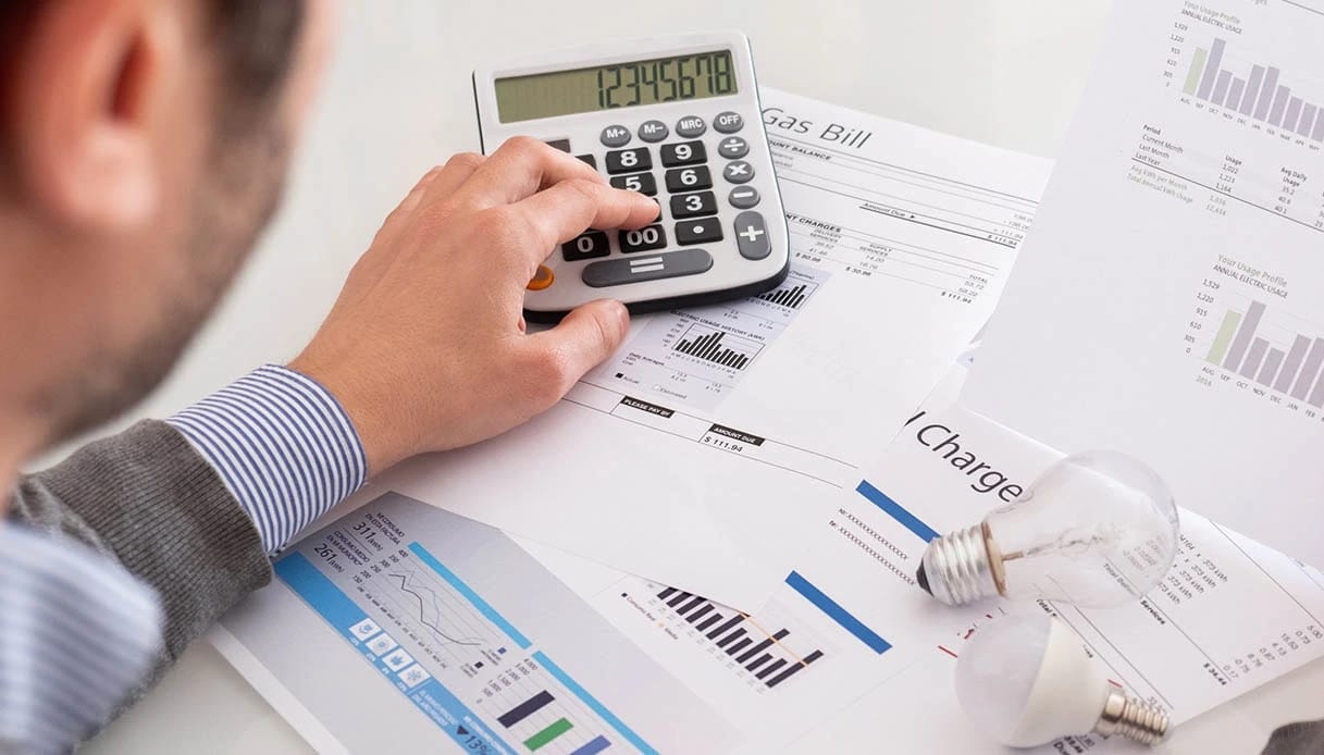 **Alt text (in Italian):**
Uomo calcola bollette energetiche con grafici e lampadine sul tavolo.